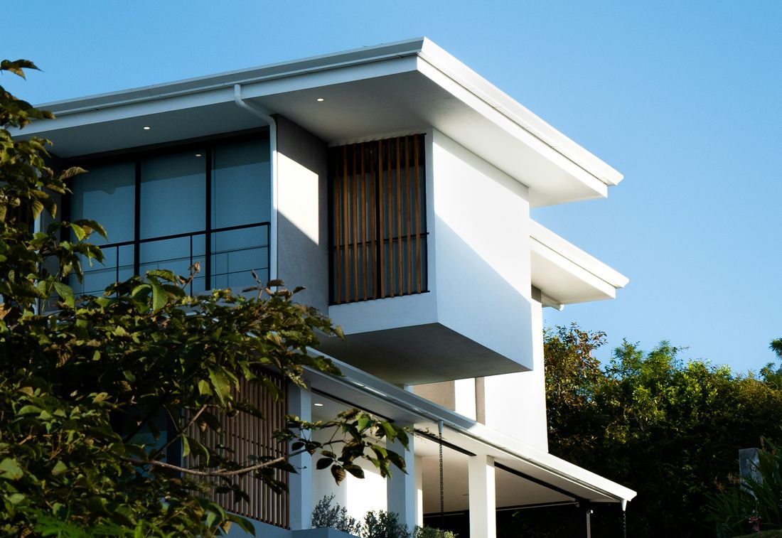A modern white house with wood slat screens and deep roof overhangs, framed by trees under a blue sky